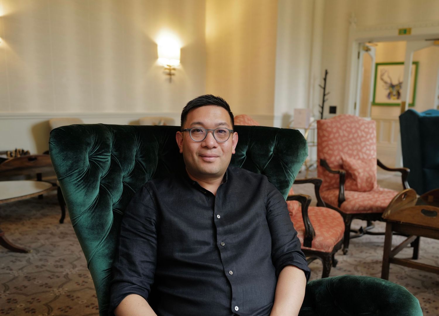 Someone wearing glasses and a black shirt sits smiling in an emerald green velvet armchair in a warmly lit room.