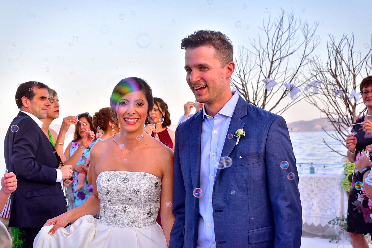 Happy newlyweds sharing a laugh during outdoor celebration with bubbles and coastal scenery.