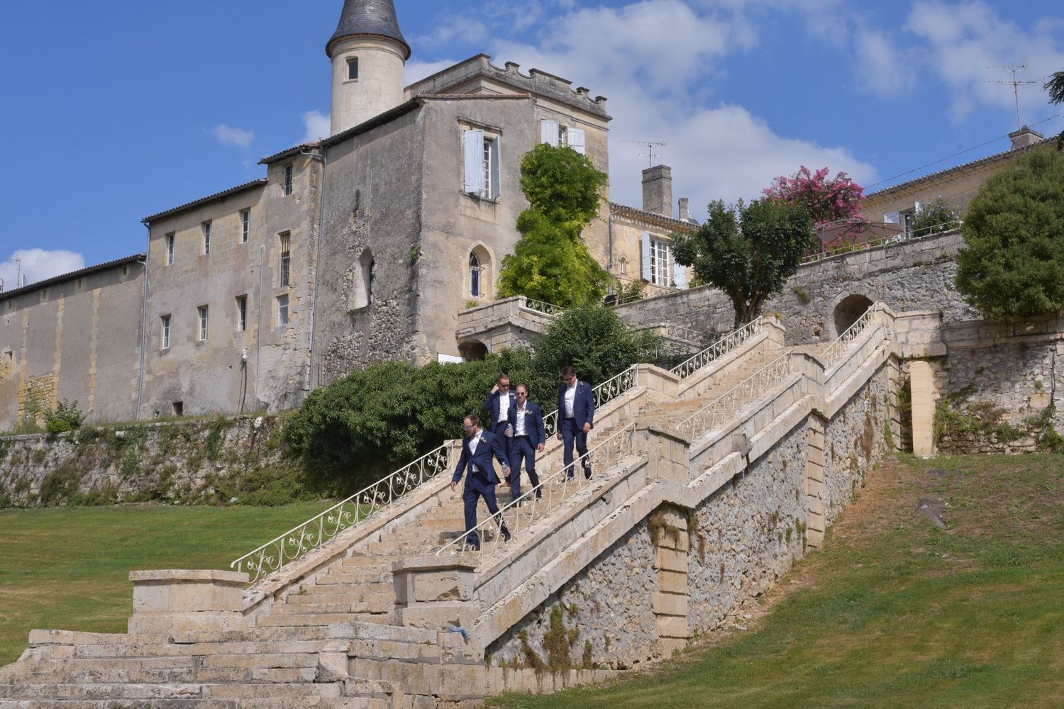Tourists descend stone staircase at historic stone castle with tower and fortress walls on grassy hillside.