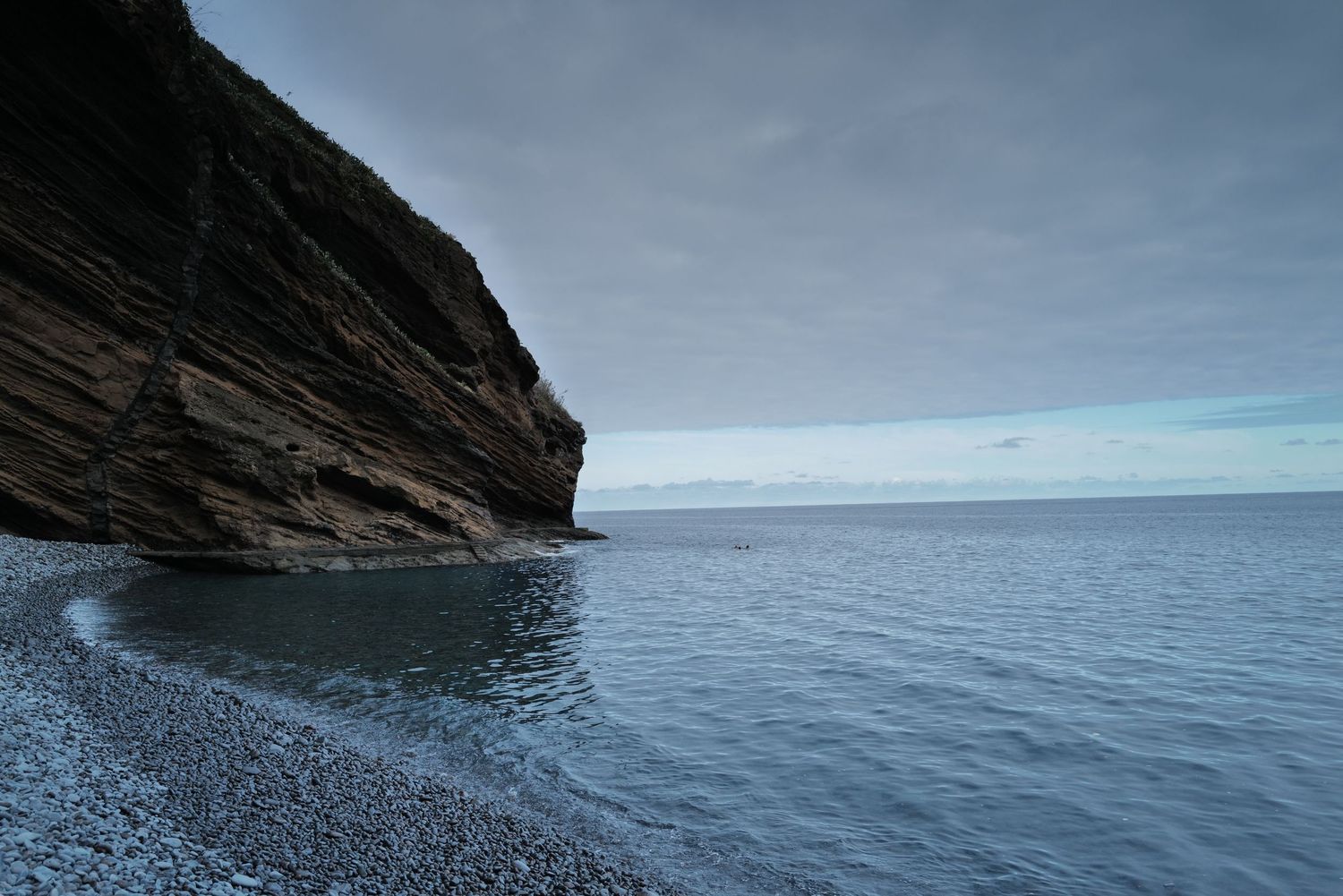 Panoramic view of rocky coastal cliffs meeting calm blue ocean waters under overcast skies.