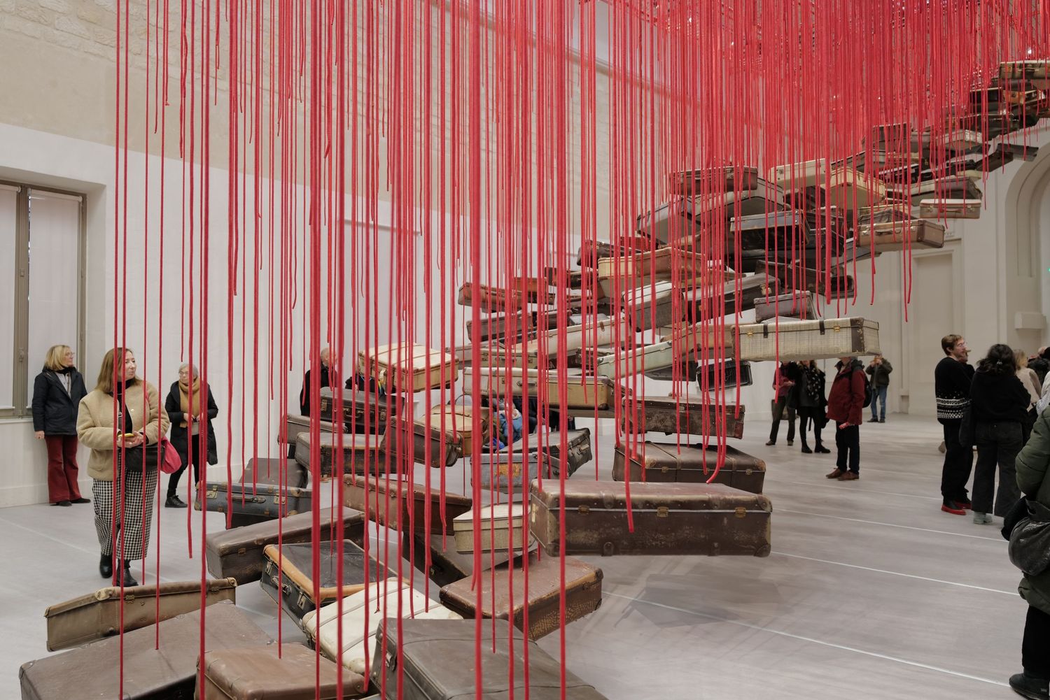 Red vertical strings form a striking art installation in a white gallery space with visitors.