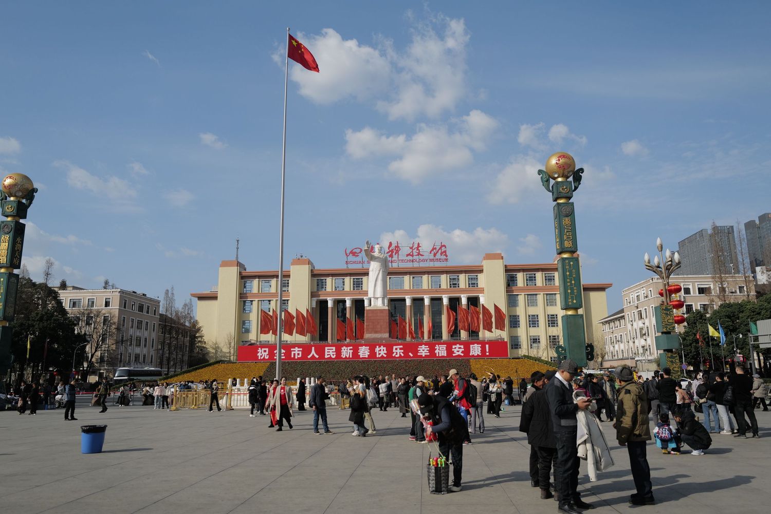 Large public square in China with red flag and government building flanked by ornate pillars.