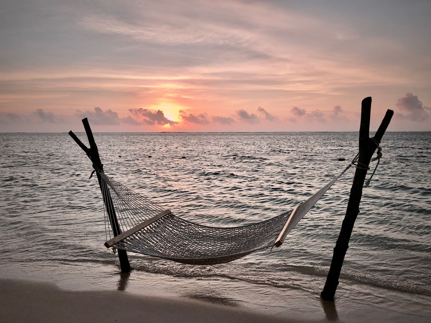 Empty hammock strung between wooden posts on a beach at sunset over calm ocean waters.