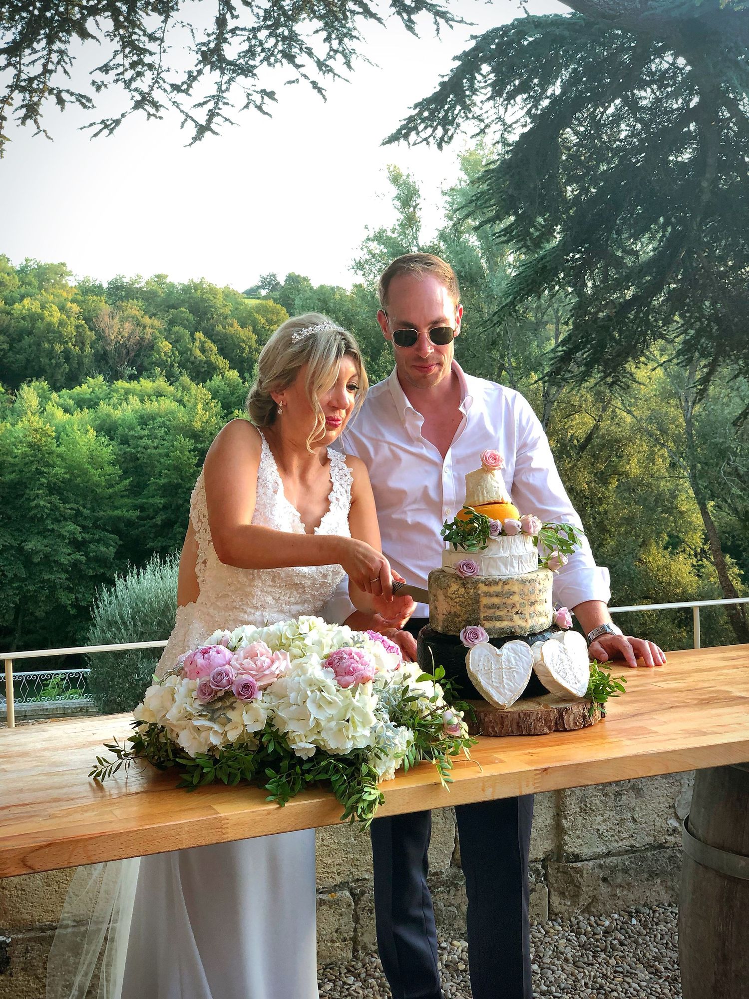 A couple cuts their wedding cake together outdoors on a wooden table decorated with pink and white floral arrangements.
