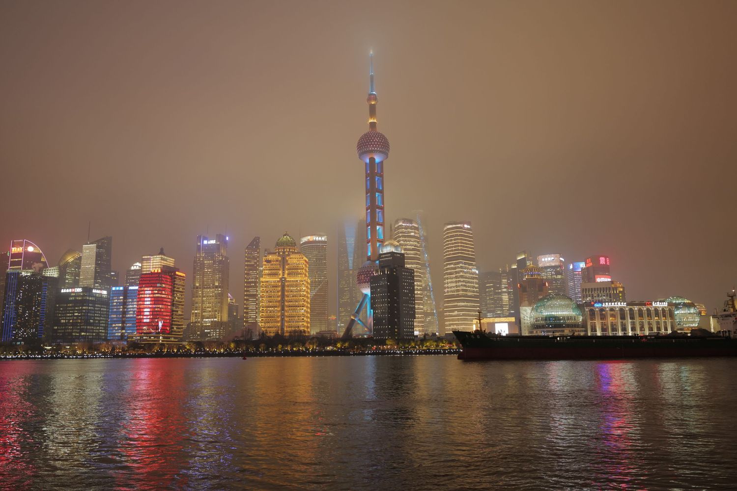 Misty night view of Shanghai's modern skyline with illuminated skyscrapers reflecting on water.
