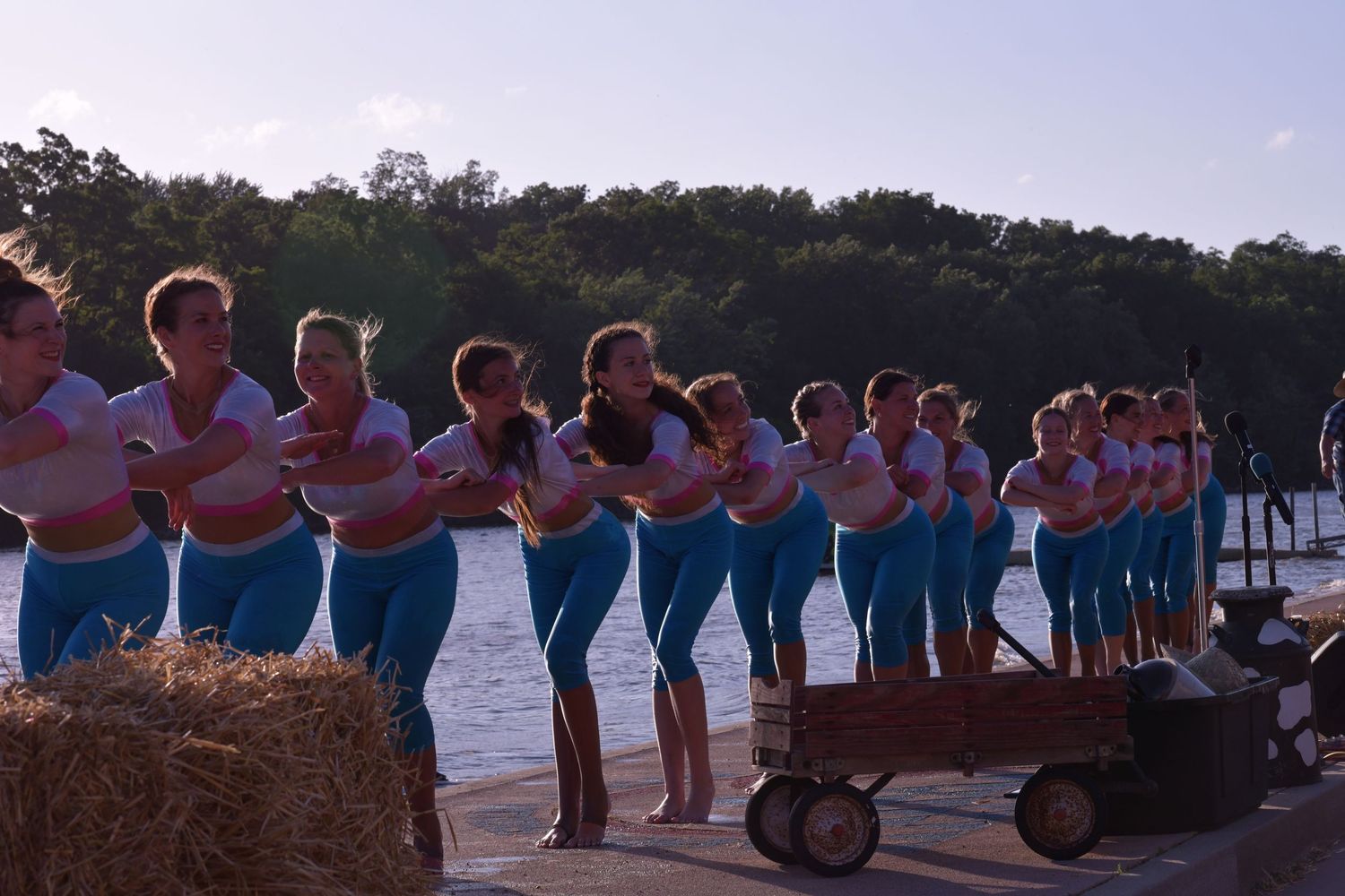 Dance performers line up on a dock at sunset wearing matching turquoise outfits against a lakeside backdrop.