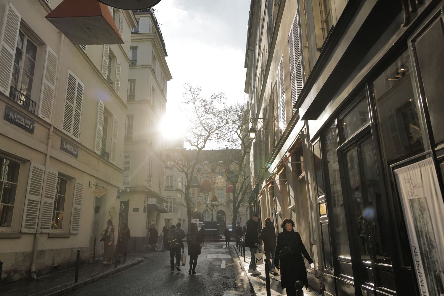 Sunlit cobblestone street in Paris with historic buildings and pedestrians walking.