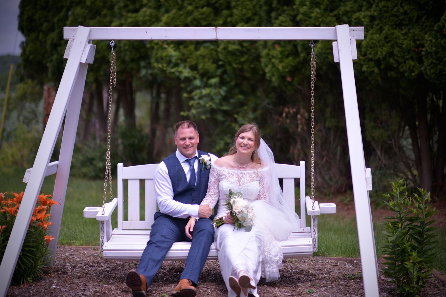 A couple sits together on a white wooden porch swing in a garden setting with greenery in the background.