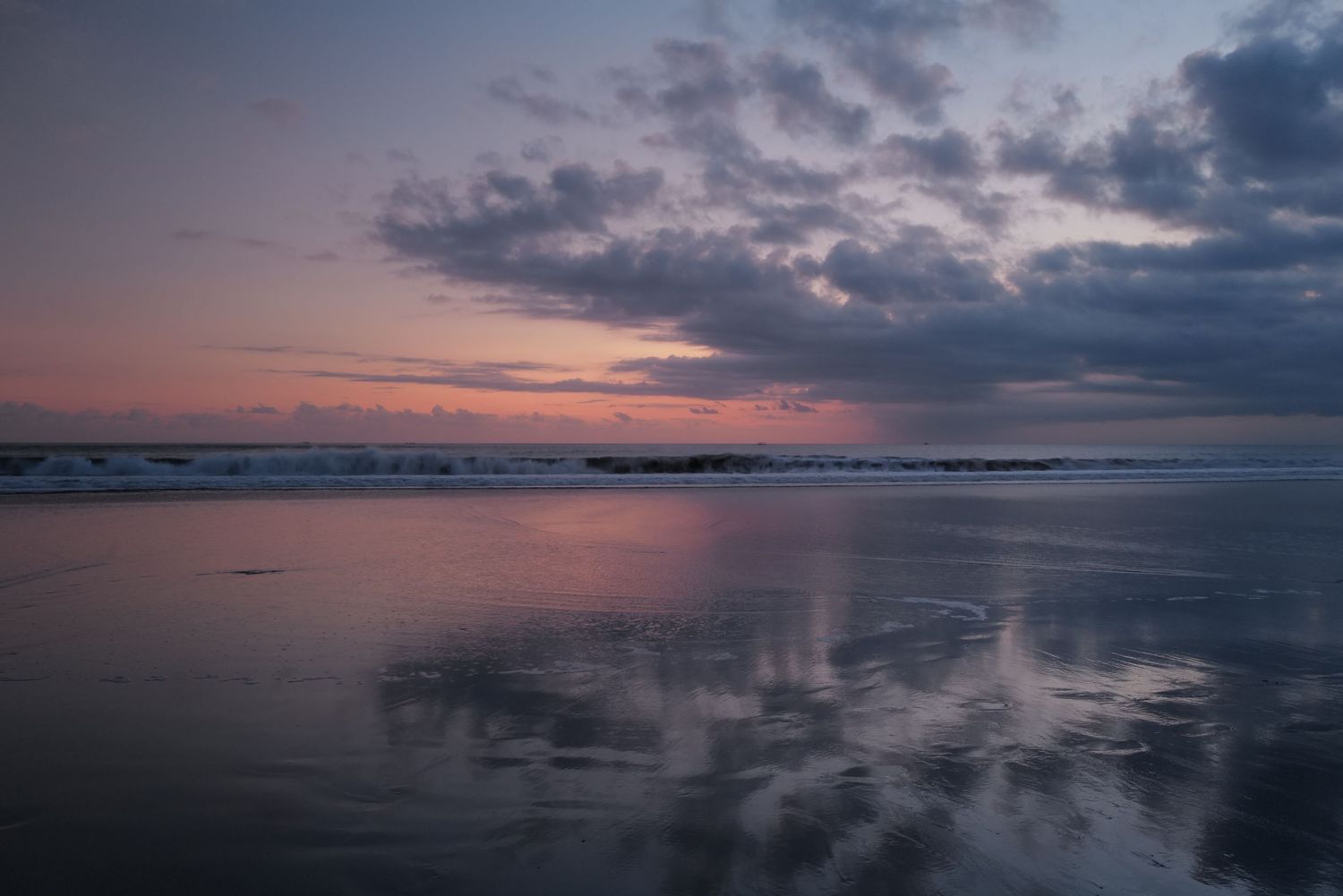Dramatic pink and purple sunset sky reflected in calm ocean waters with dark clouds.