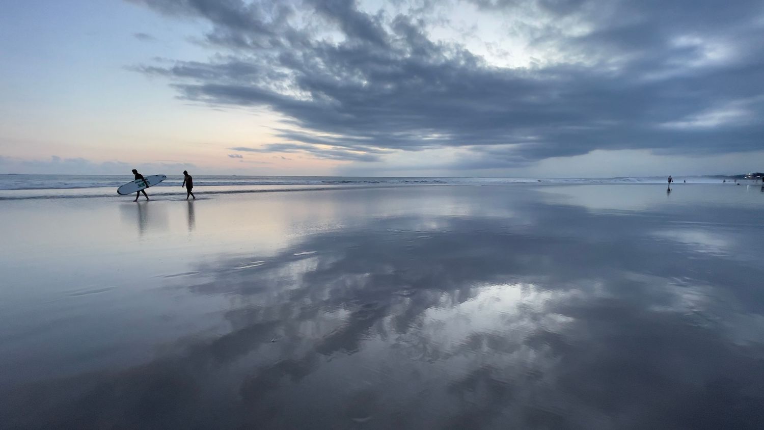 Silhouettes of surfers on reflective beach at sunset with dramatic cloudy sky.