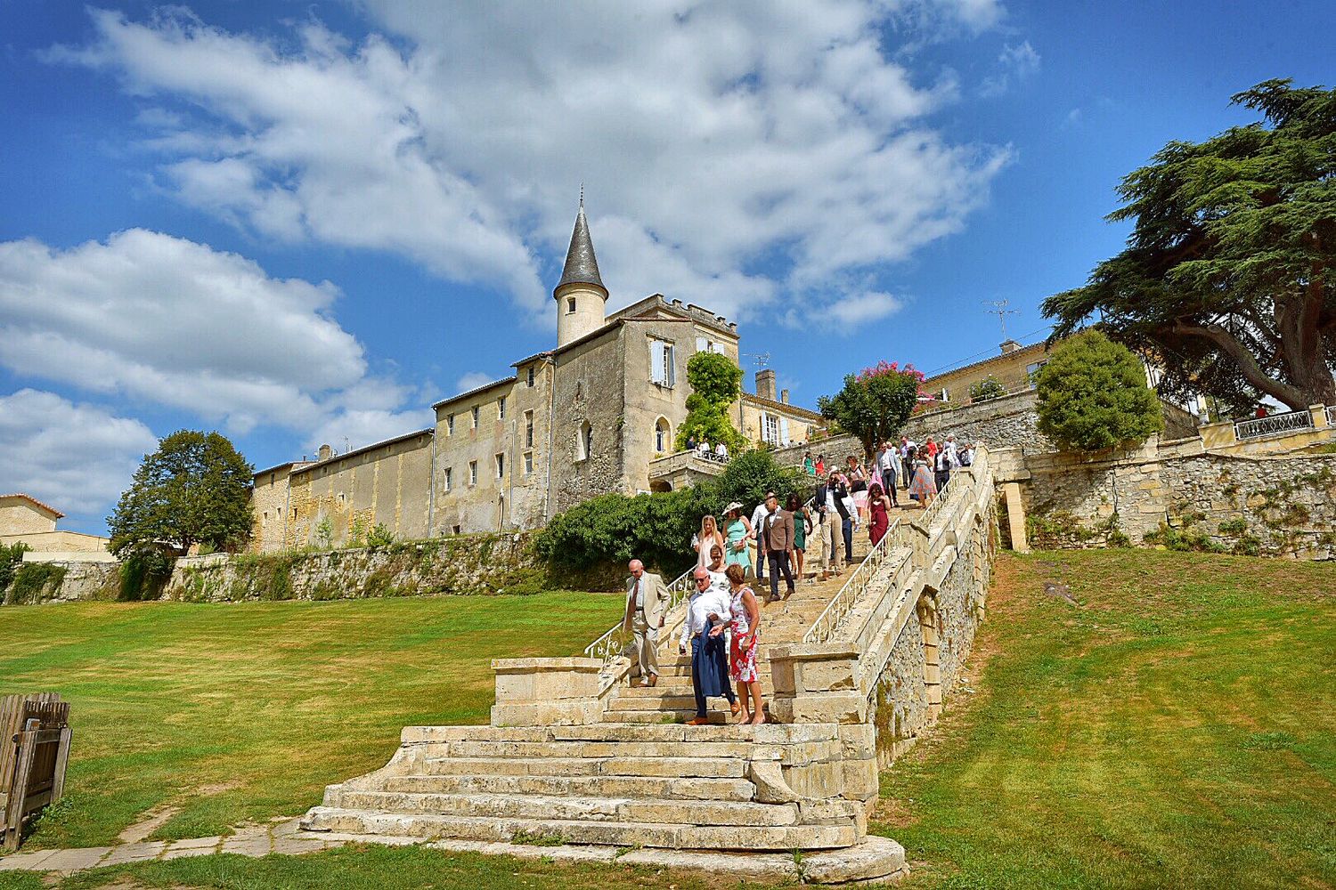 Visitors climb stone steps leading to a historic chateau under blue skies and white clouds in France.
