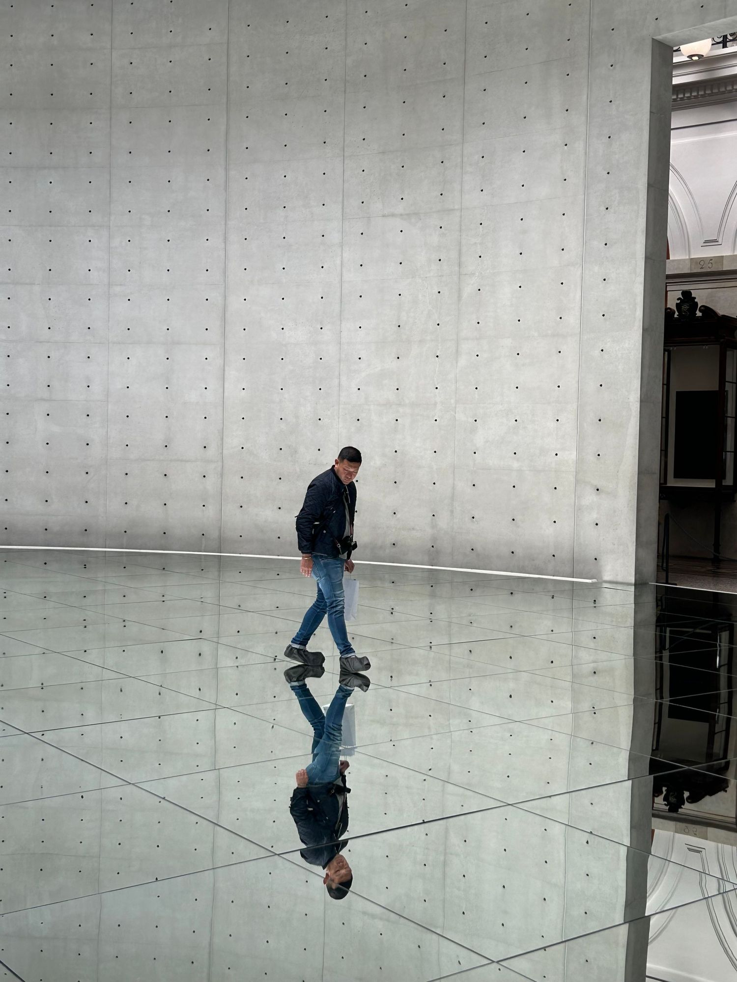 Minimalist white gallery space with reflective floor showing a person walking and their mirrored reflection.