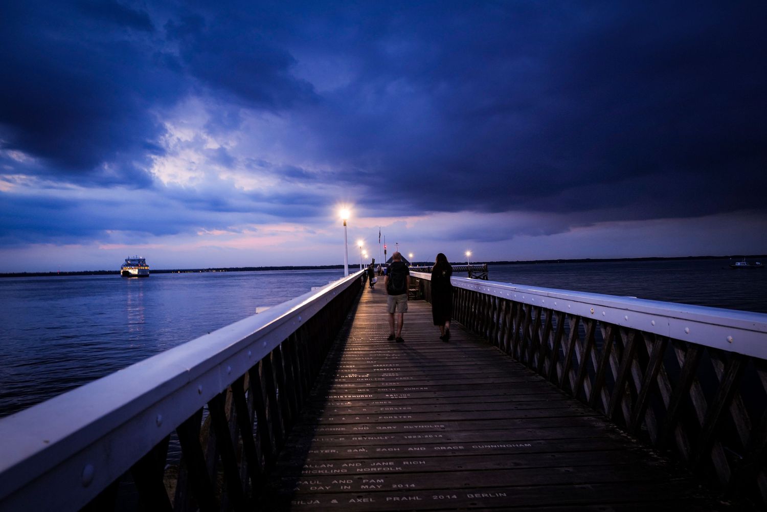 Dramatic dark boardwalk at twilight with lightning strikes illuminating stormy clouds over a serene harbor.