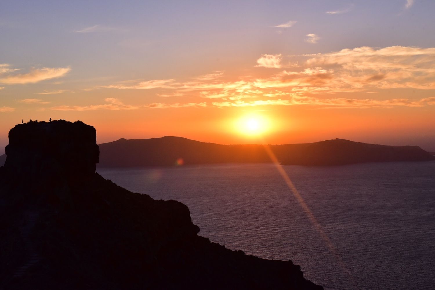 Golden sunset silhouettes rocky cliffs against a glowing orange sky over distant mountain islands.