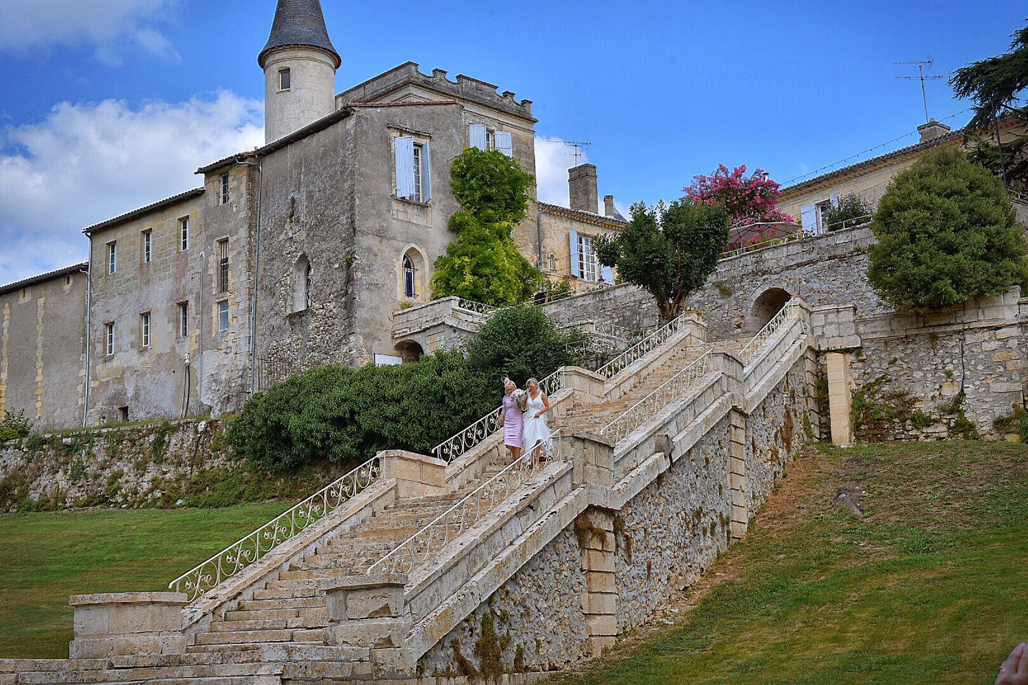 Historic stone chateau with tall turret and grand stone staircase leading up hillside under blue summer sky.