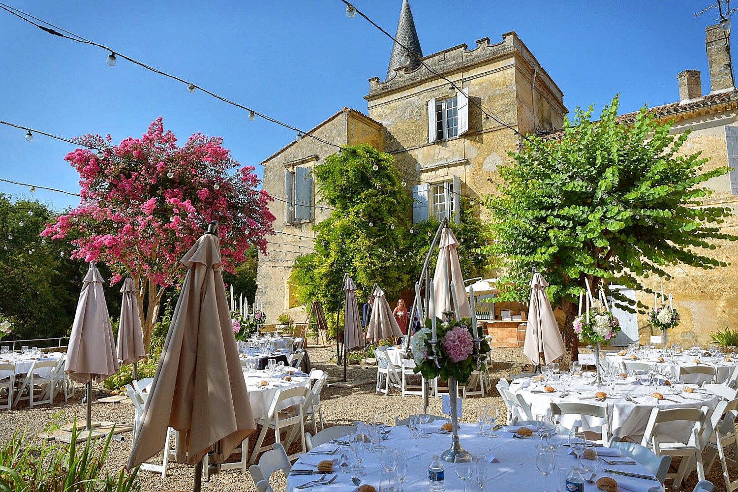 Elegant outdoor wedding reception setup with white tables in a French chateau courtyard featuring pink flowering trees.