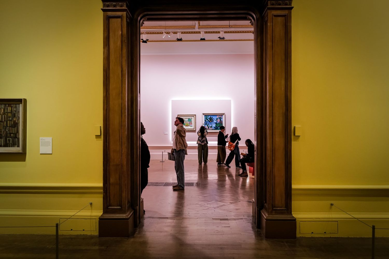Museum gallery visitors viewing art through a wooden doorframe in a yellow-walled exhibition space.