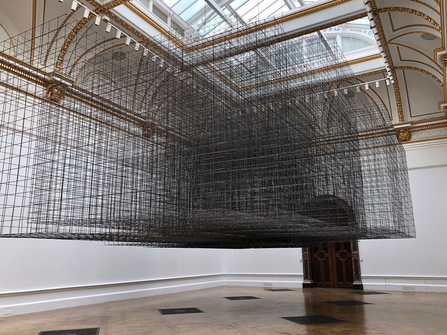 Modern art installation featuring suspended wire mesh structures under a glass ceiling.