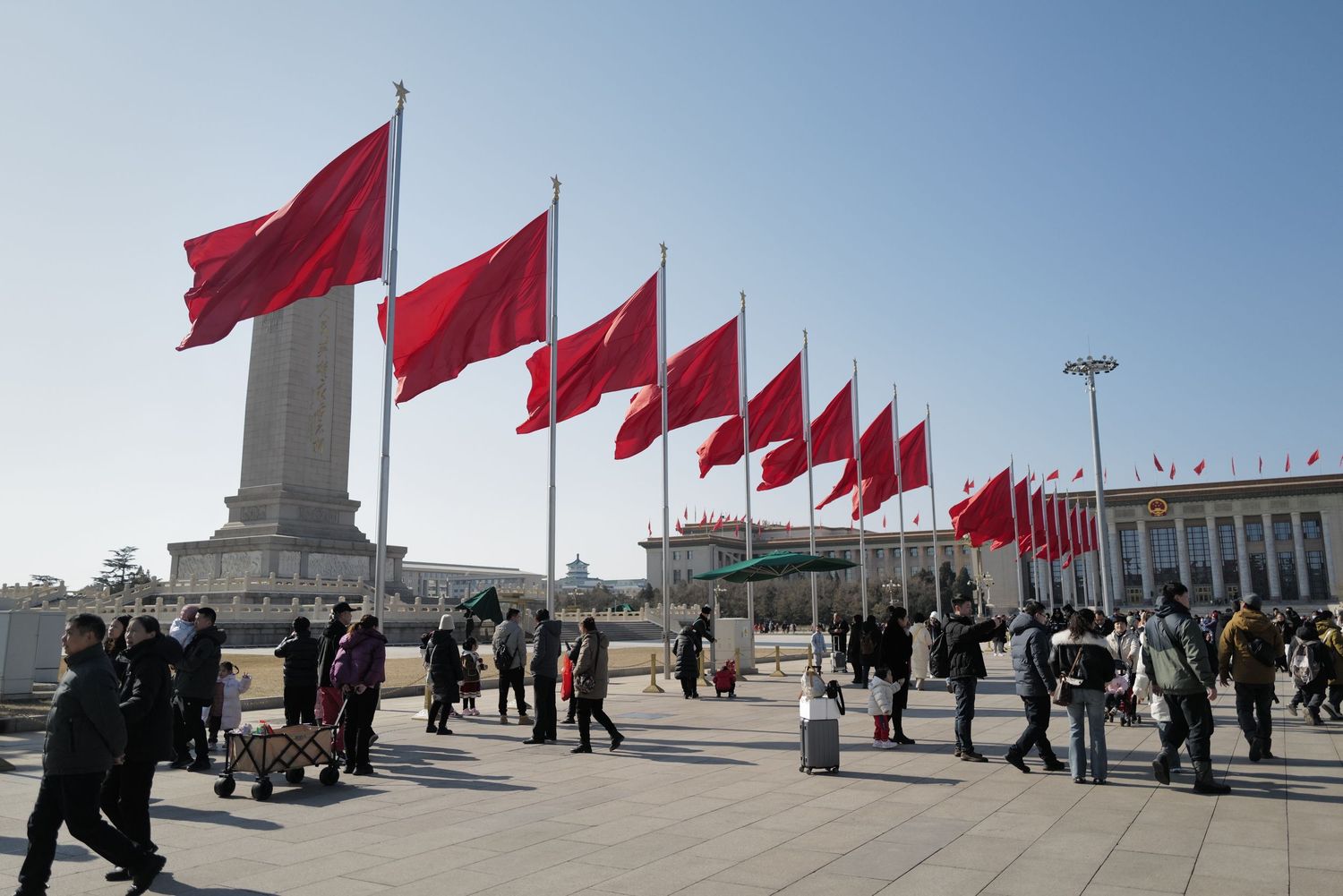 Row of red Chinese flags flying in Tiananmen Square with crowds gathered below.