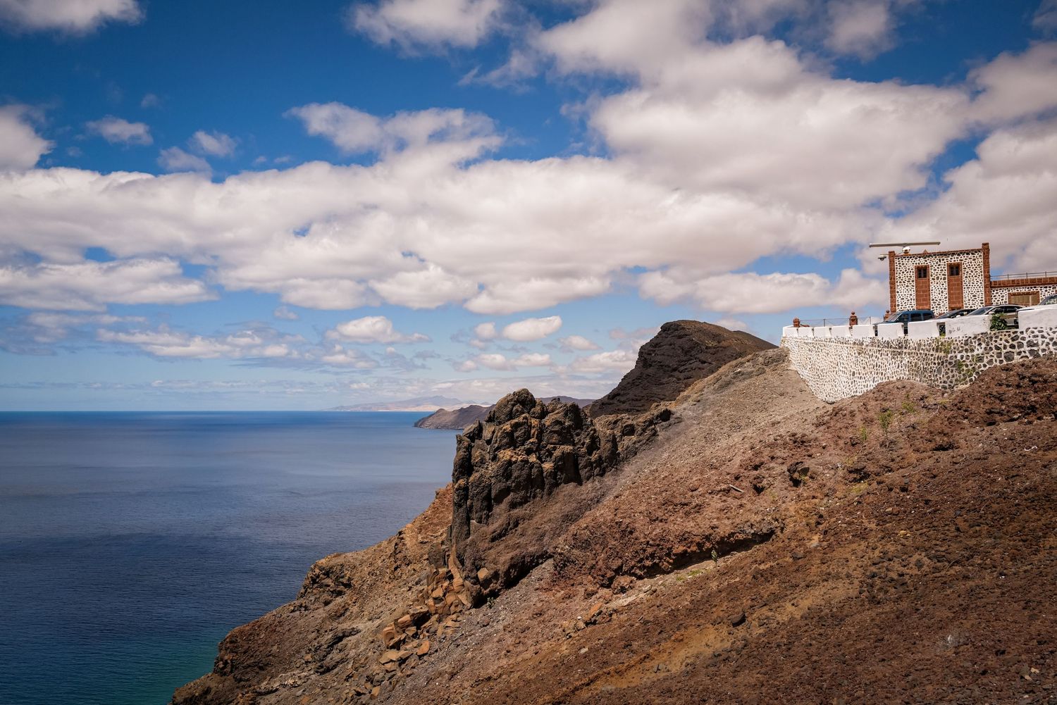 Small white building perched on rocky coastal cliffs overlooking a vast blue ocean.