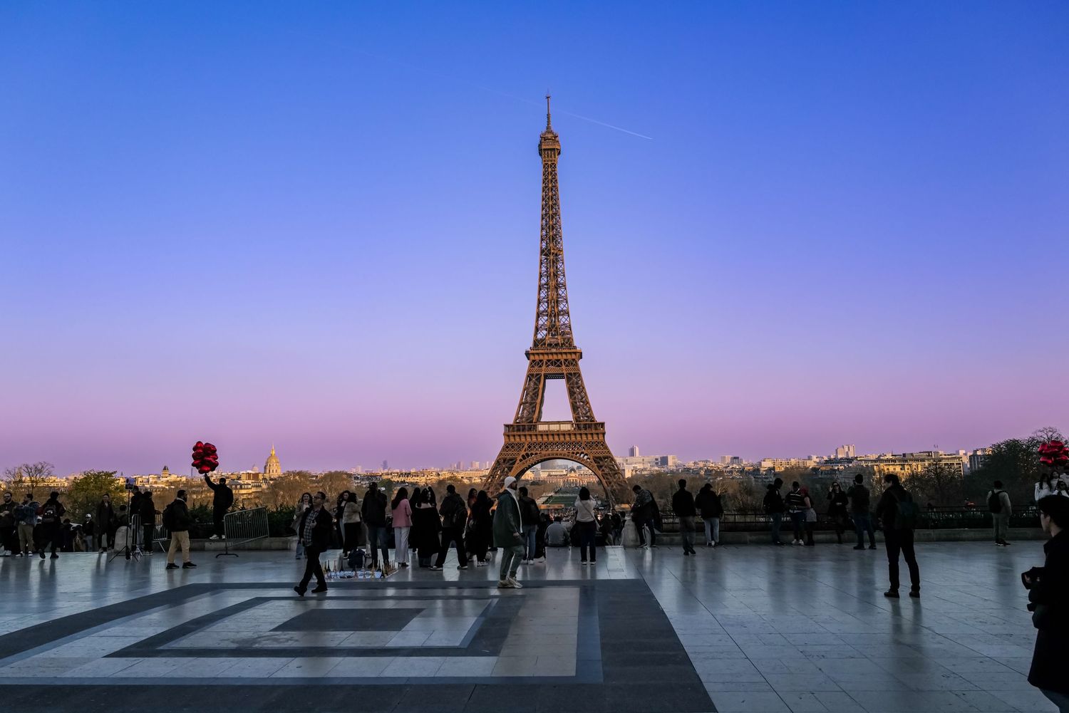 Silhouettes of people gathering at Trocadero plaza with Eiffel Tower visible against twilight sky.