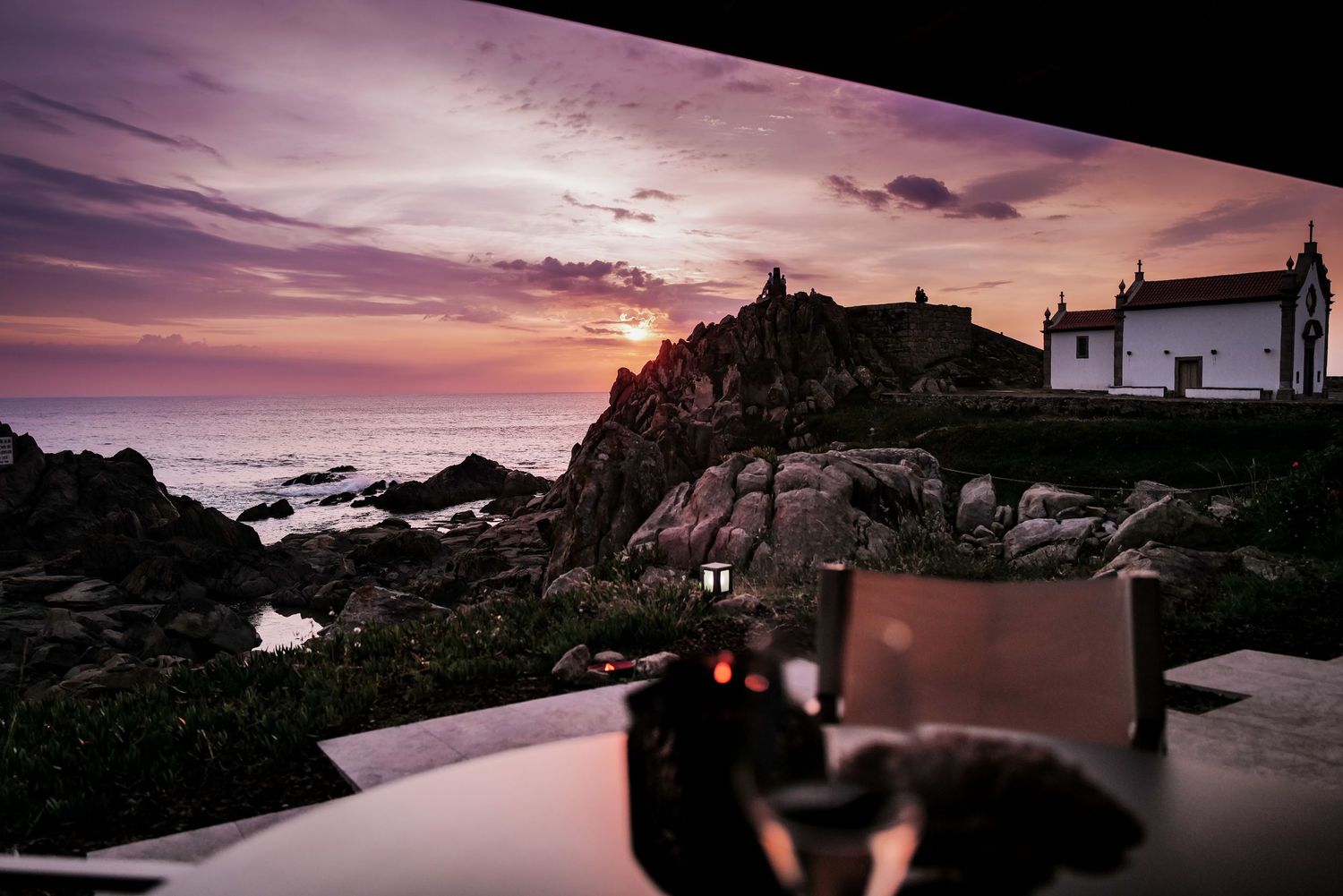 Dramatic sunset sky over coastal buildings perched on rocky cliffs with camera in foreground.
