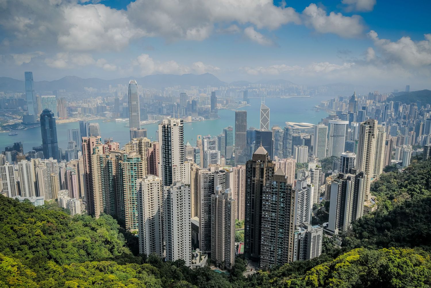Hong Kong's iconic skyline viewed from Victoria Peak with lush green mountains in foreground.