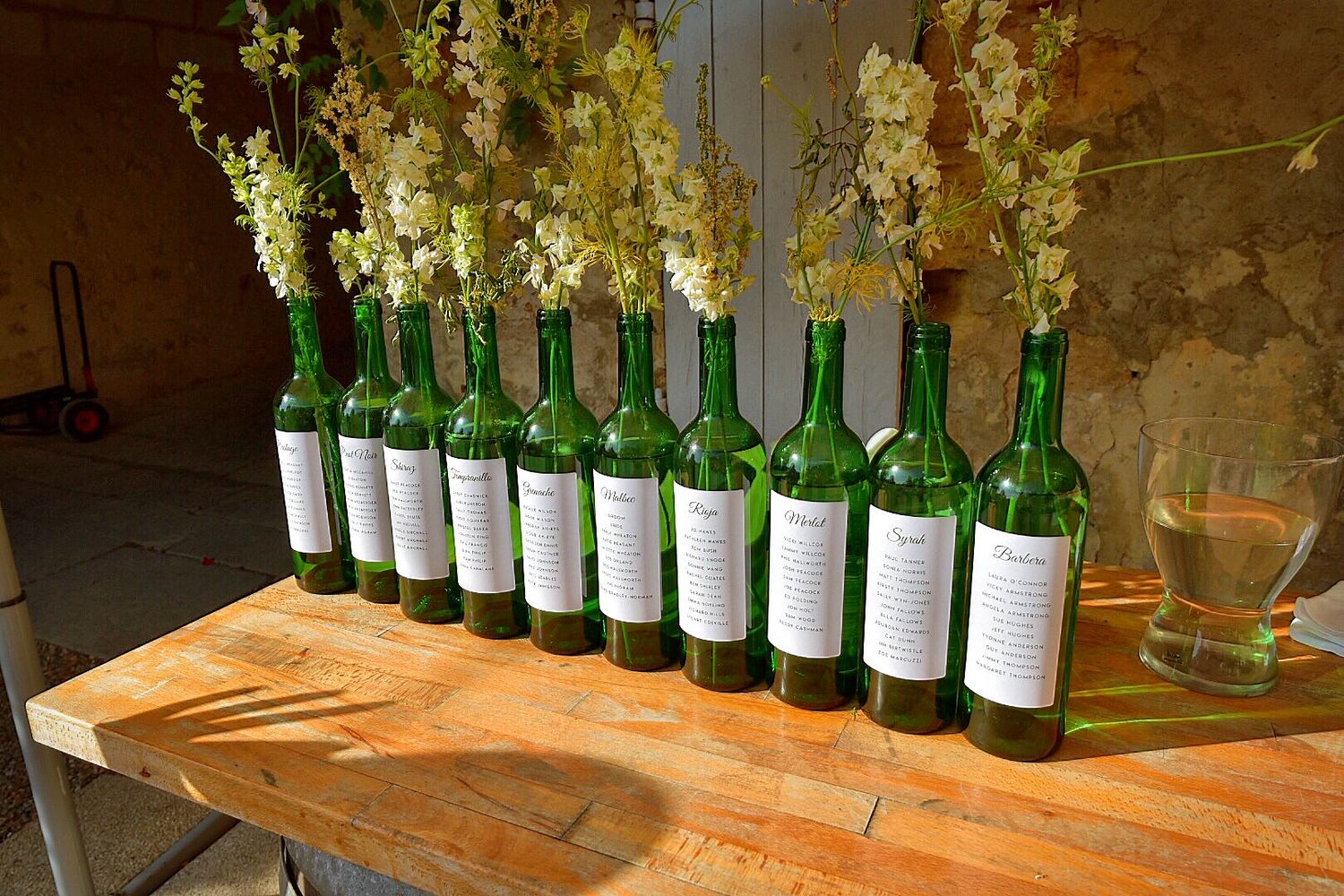 A row of green wine bottles with white labels lined up on a wooden table, each topped with delicate white flowers.