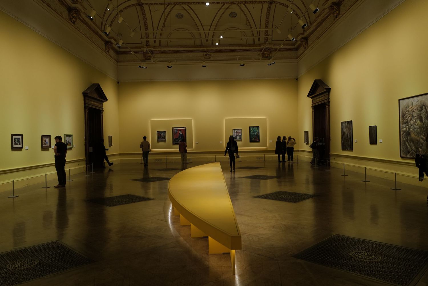 Gallery visitors admire artwork displayed in a dimly lit museum exhibition space with curved architectural elements.