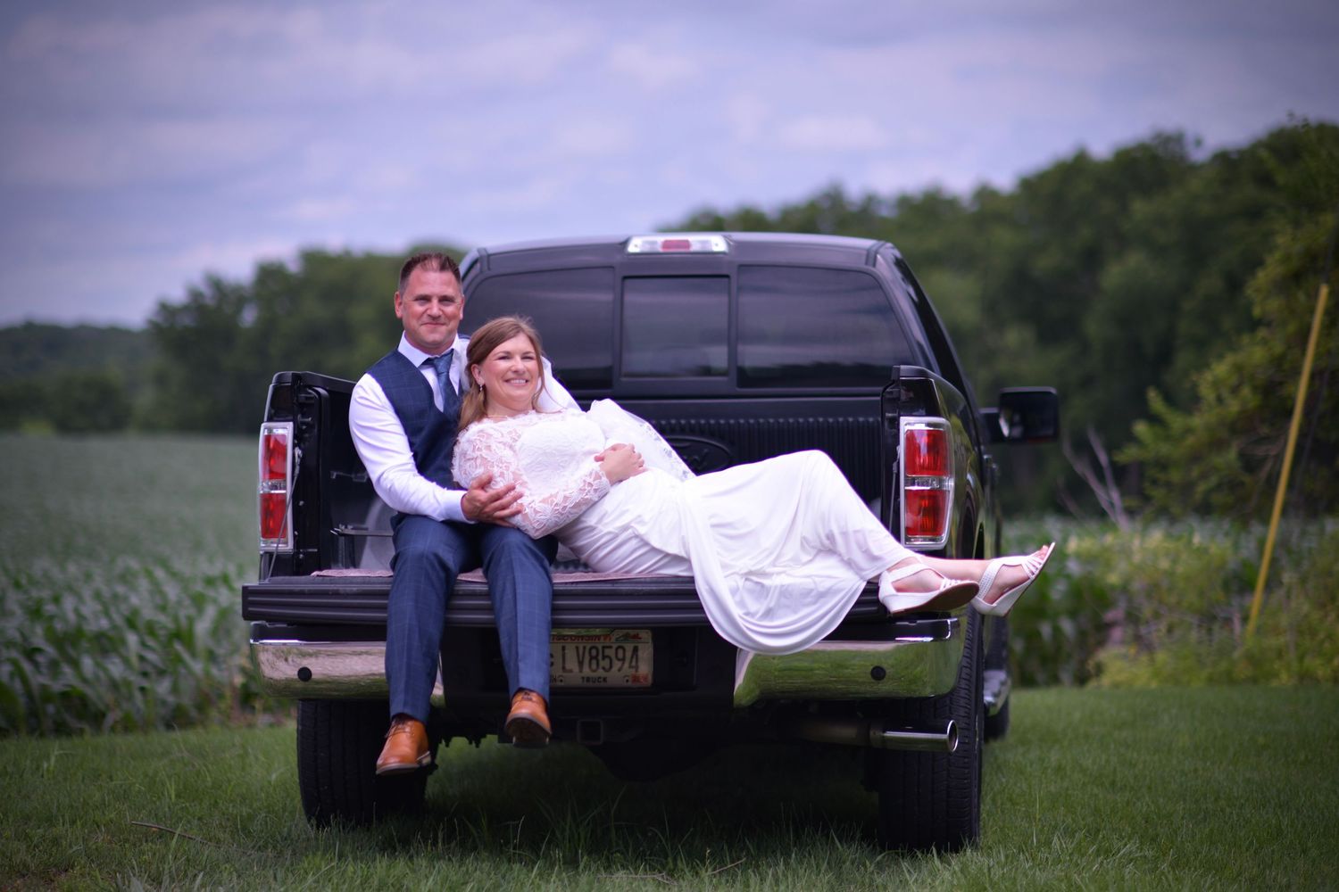 A couple lying together in the back of a pickup truck on a grassy field at sunset.