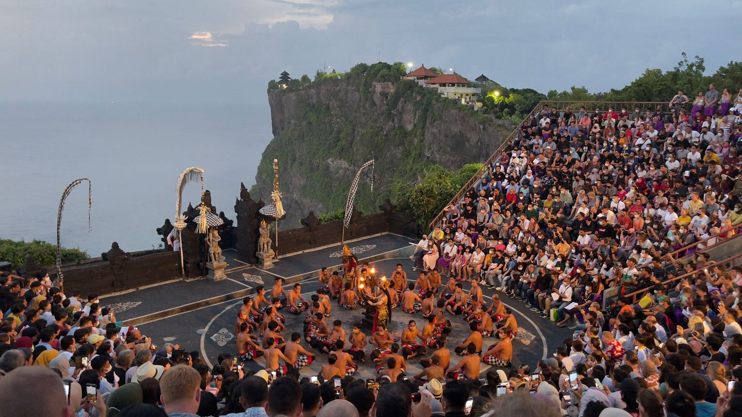 Large crowd gathered at clifftop amphitheater overlooking ocean for cultural performance in Bali.