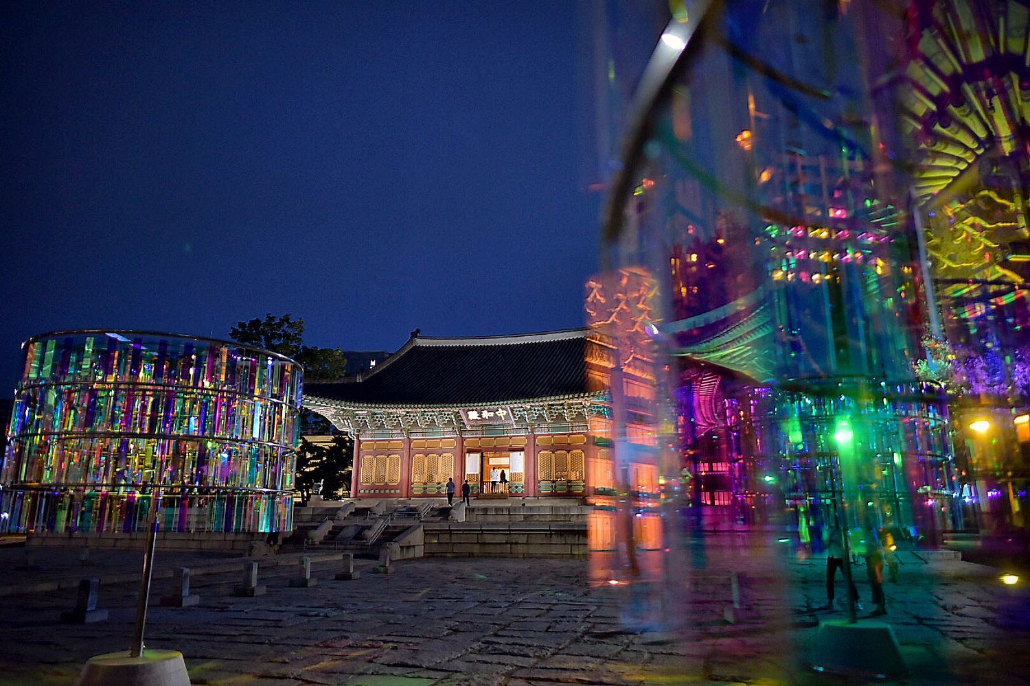 Colorful illuminated traditional Korean palace at night with vibrant decorative lighting and courtyard.