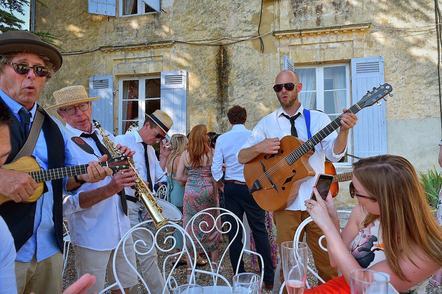 Musicians perform live outdoor music with guitars while people gather at a charming French stone building.