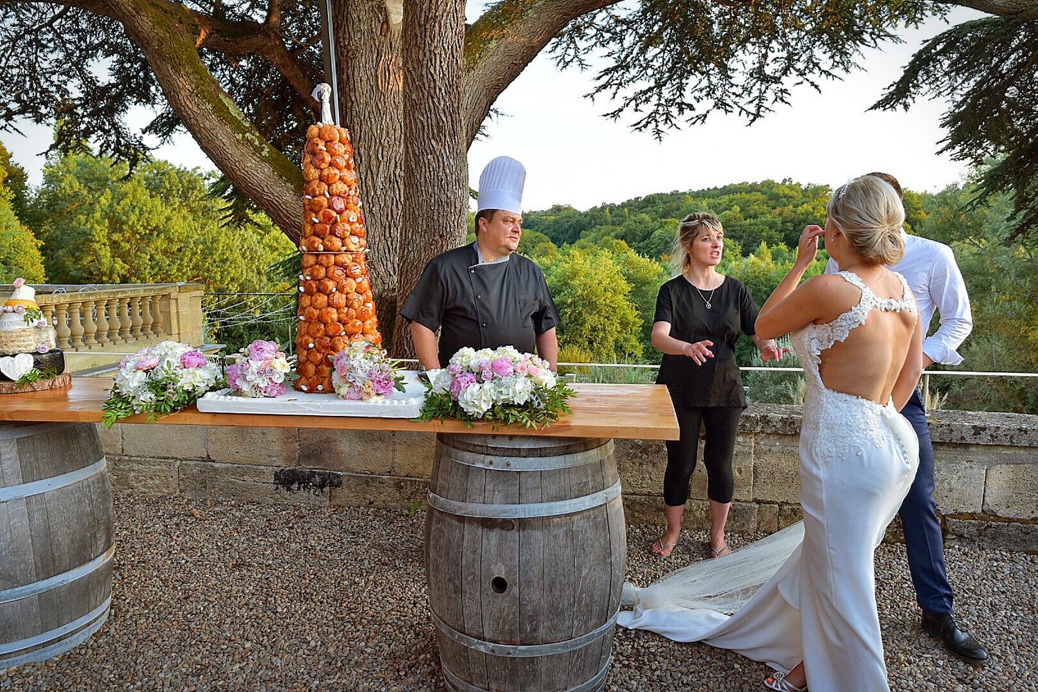 Wedding party enjoys refreshments at rustic outdoor bar setup with wine barrels and decorative fruit tower display.