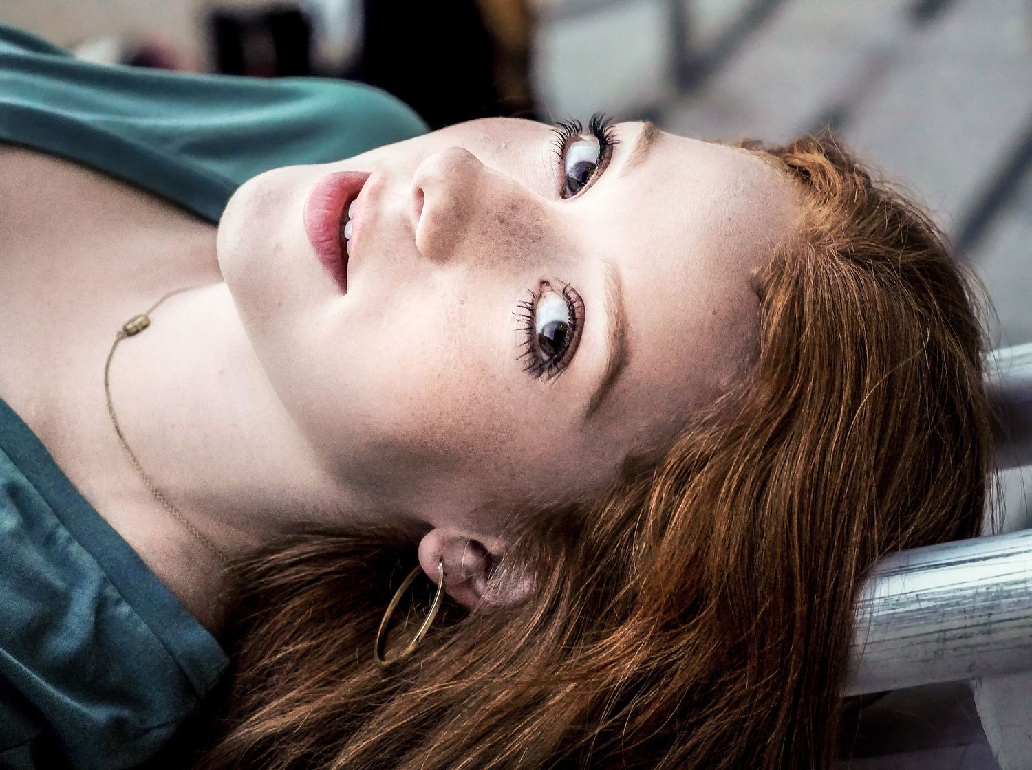 Young woman with red hair and freckles lying back, looking up with a thoughtful expression outdoors.