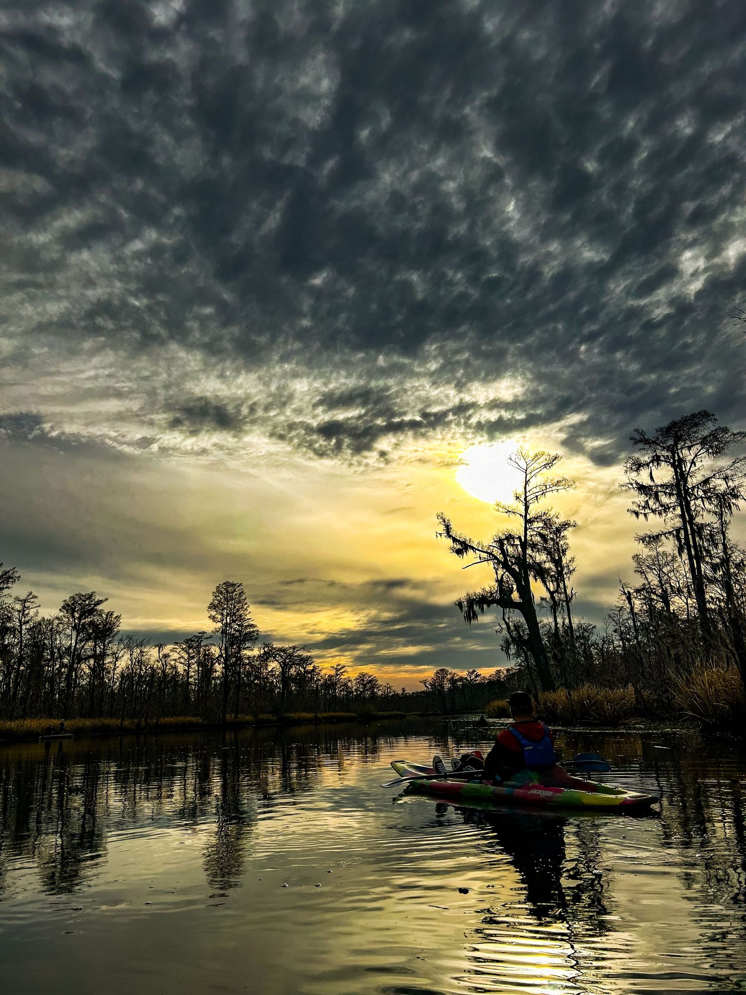 A kayaker paddling on a calm swamp at sunset under dramatic cloudy skies with cypress trees reflected in water.