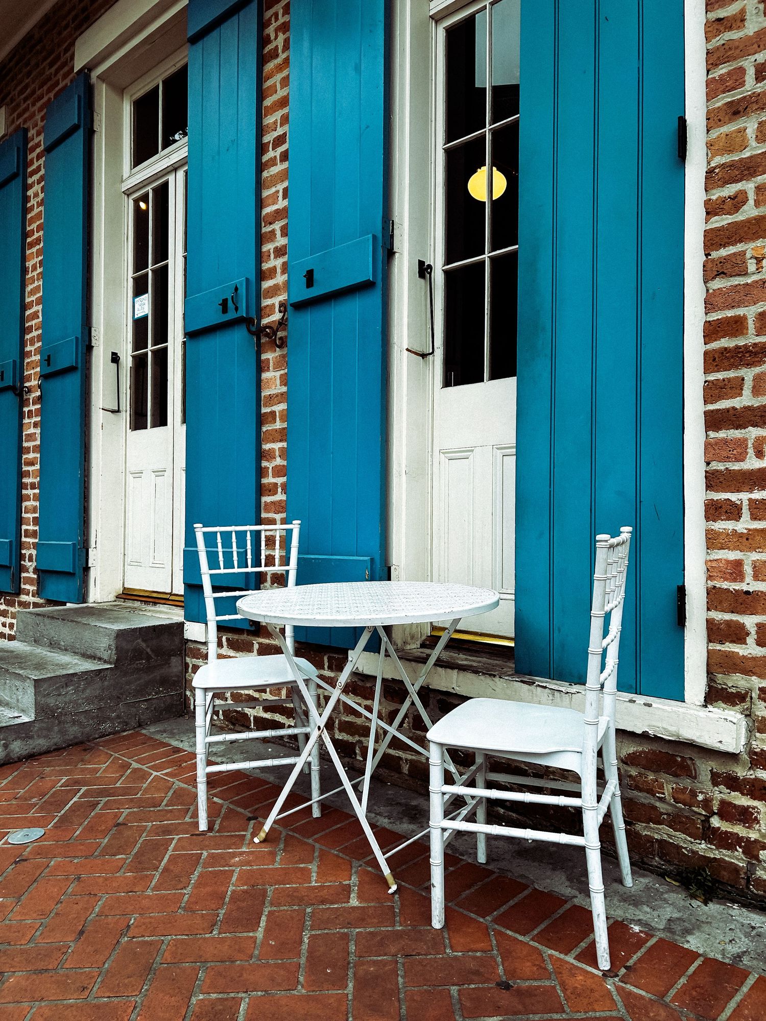 White bistro table and chairs on brick patio outside building with blue shutters and white doors.