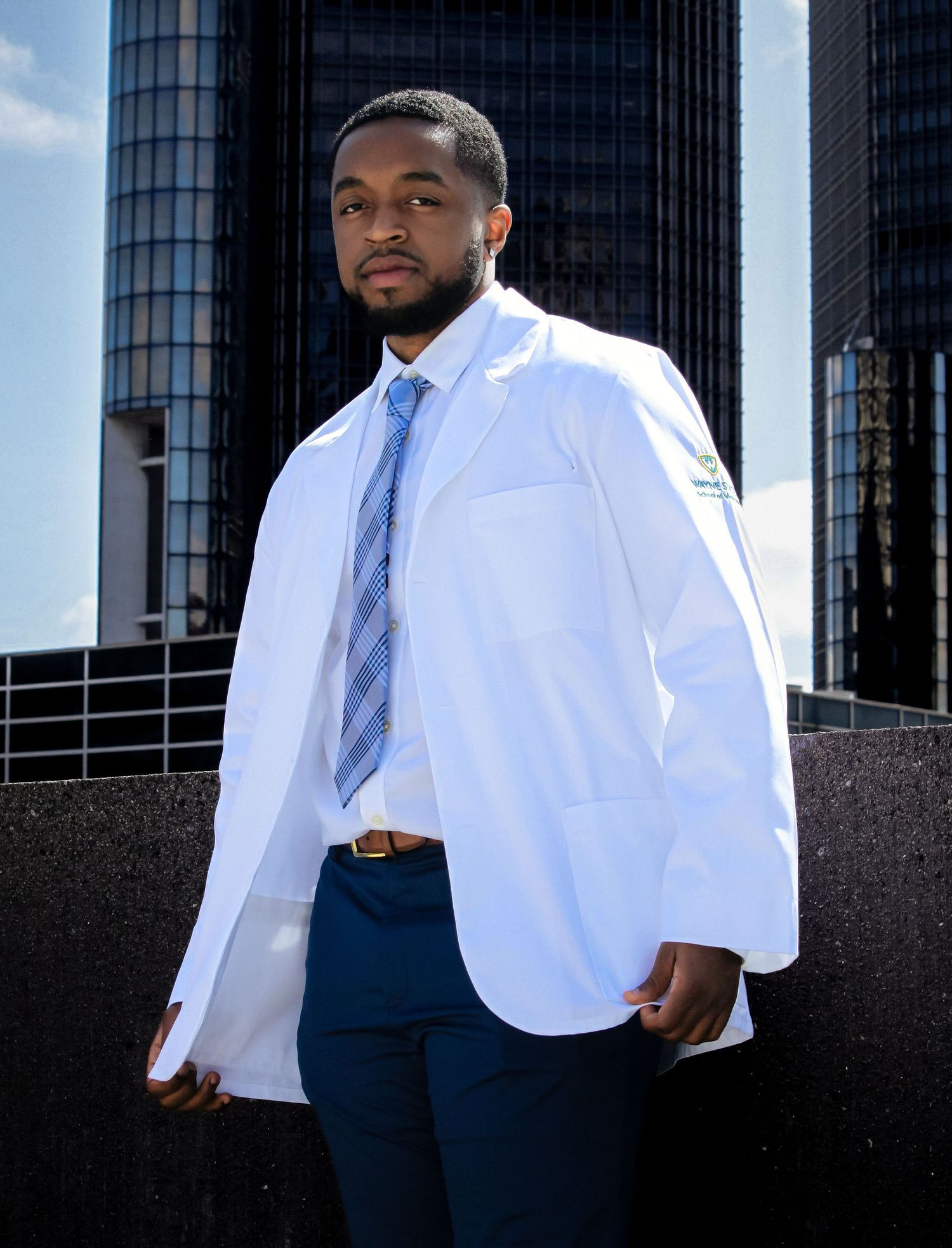 Young man in white medical coat and blue tie poses confidently outdoors with city skyscrapers in background.