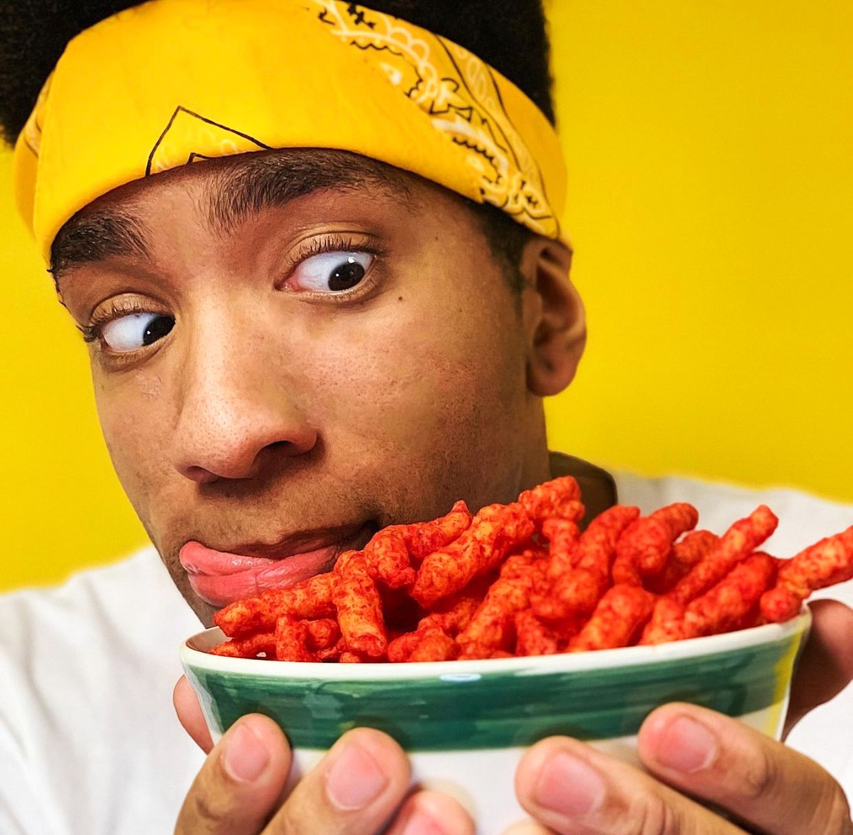 Young man with yellow bandana holding bowl of Flamin Hot Cheetos, making a funny face with tongue sticking out against yellow background.