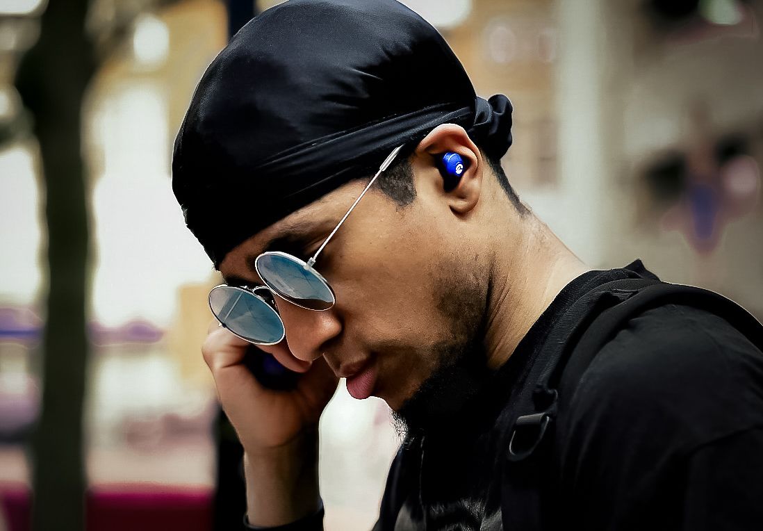 Young man wearing black durag and blue round sunglasses talking on phone in urban setting.