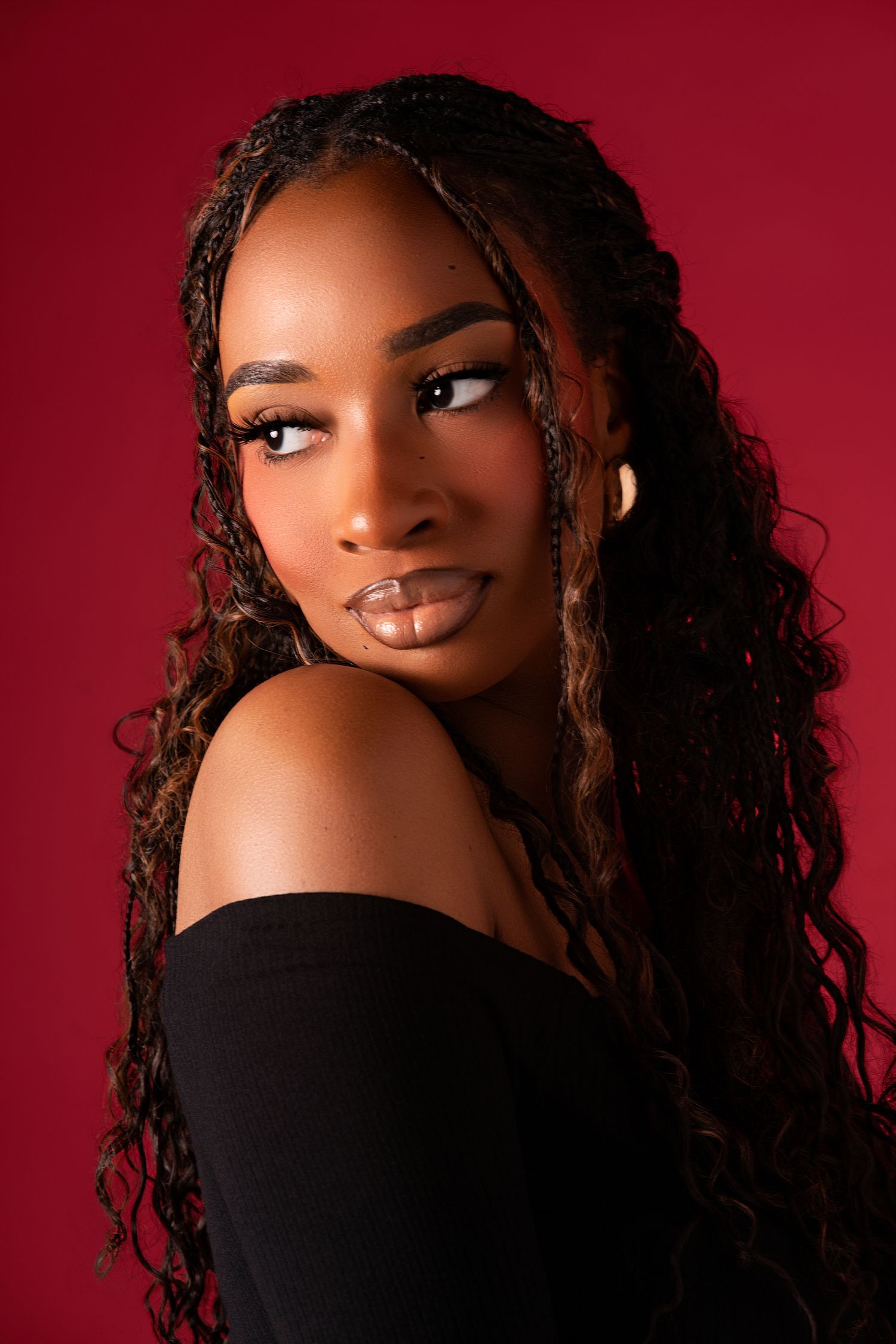 Young Black woman with long braided hair poses in black off-shoulder top against red background.