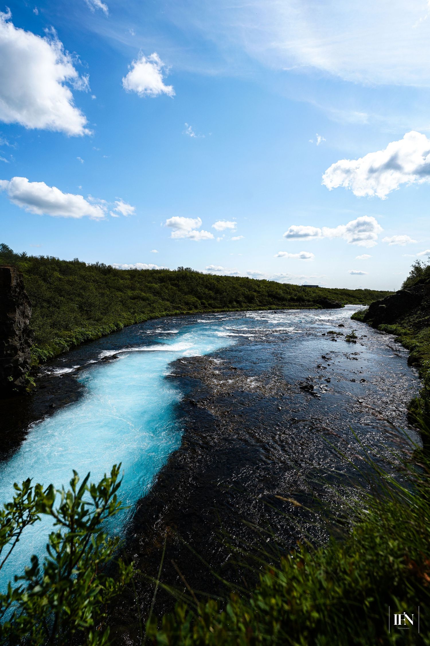 Vivid turquoise glacial river flowing through lush green Iceland landscape under bright blue sky with white clouds.