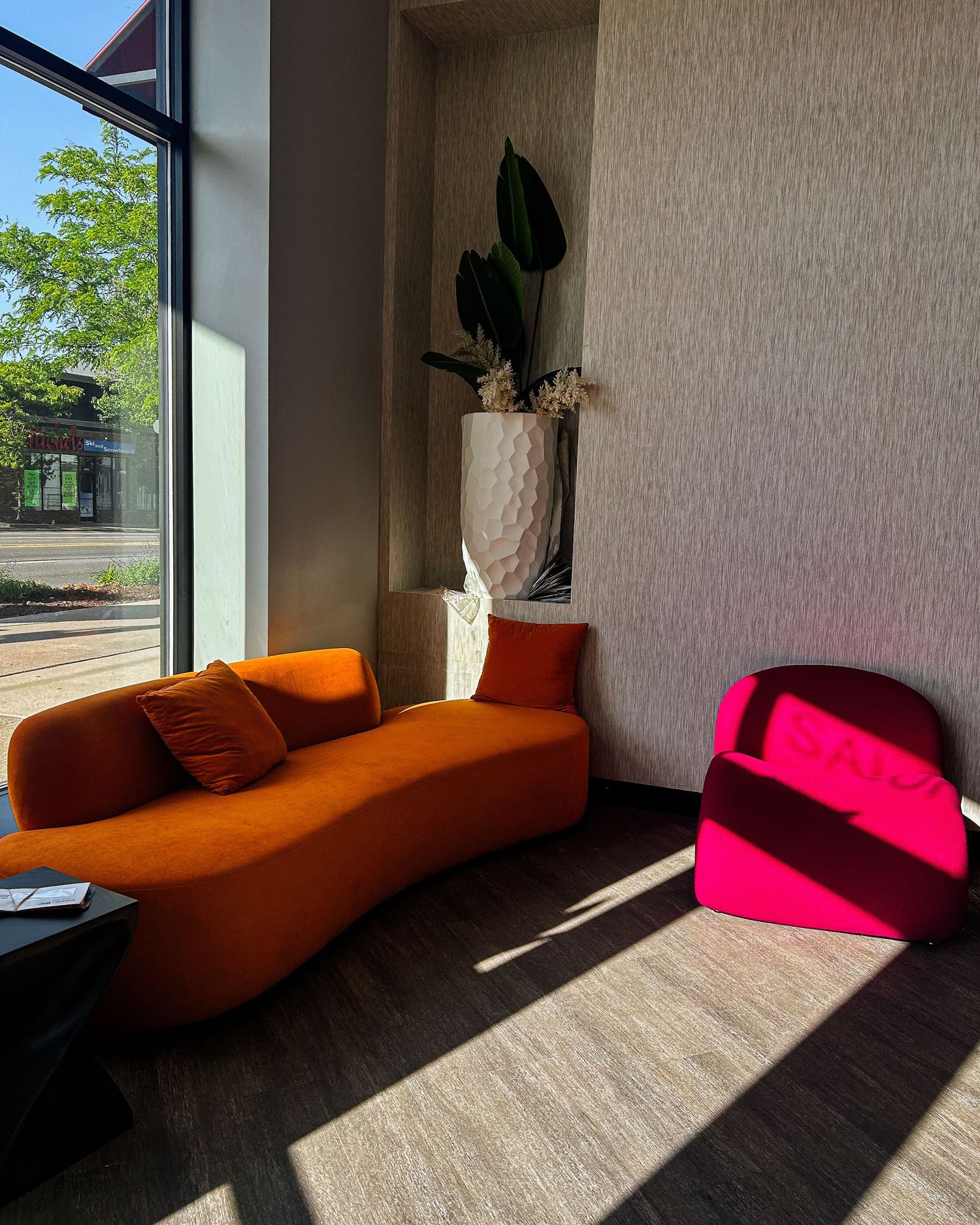 Modern lobby with curved orange sofa and hot pink chair bathed in sunlight near floor-to-ceiling windows.