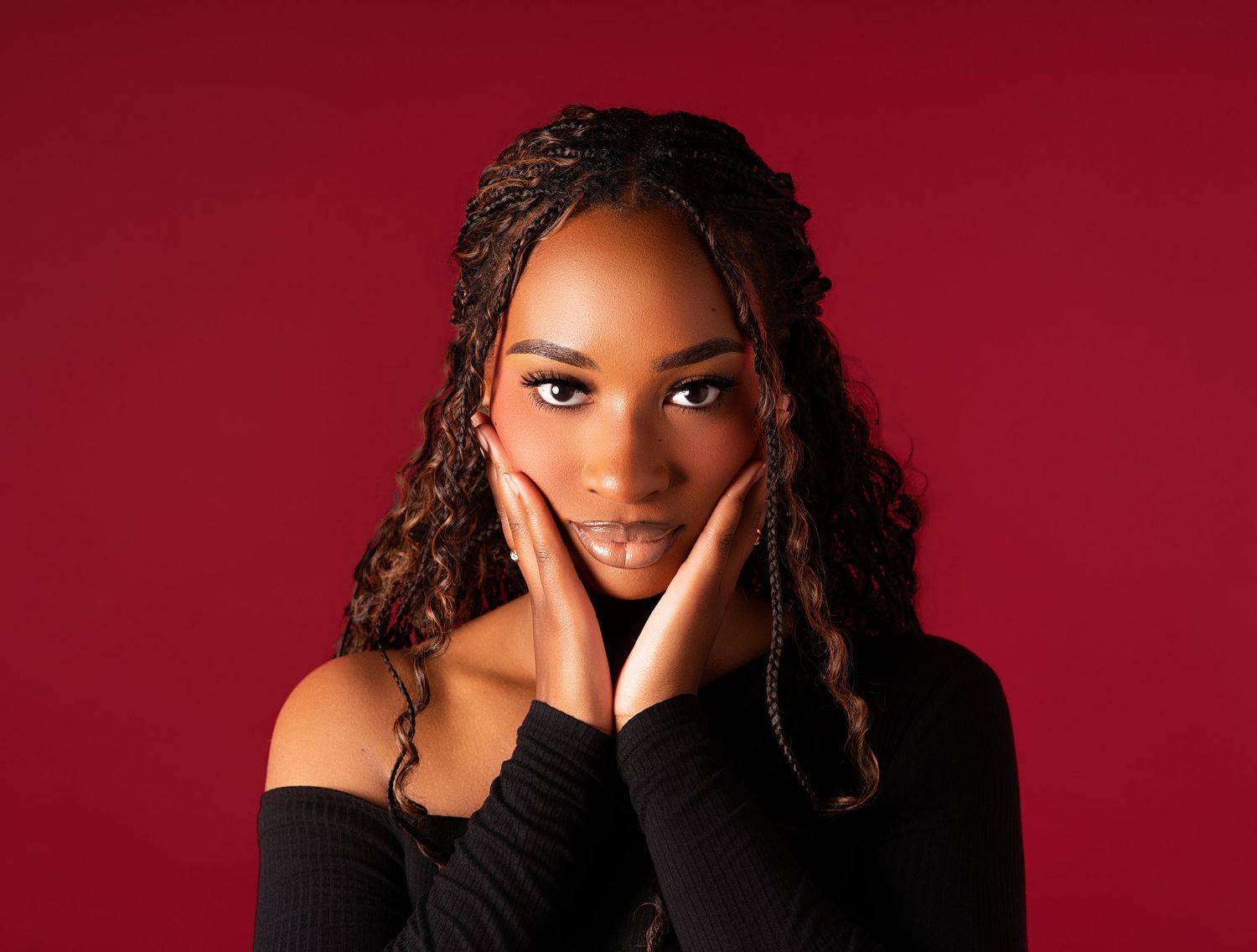 Young Black woman with braided curly hair poses gracefully against a red background in a black off-shoulder top.