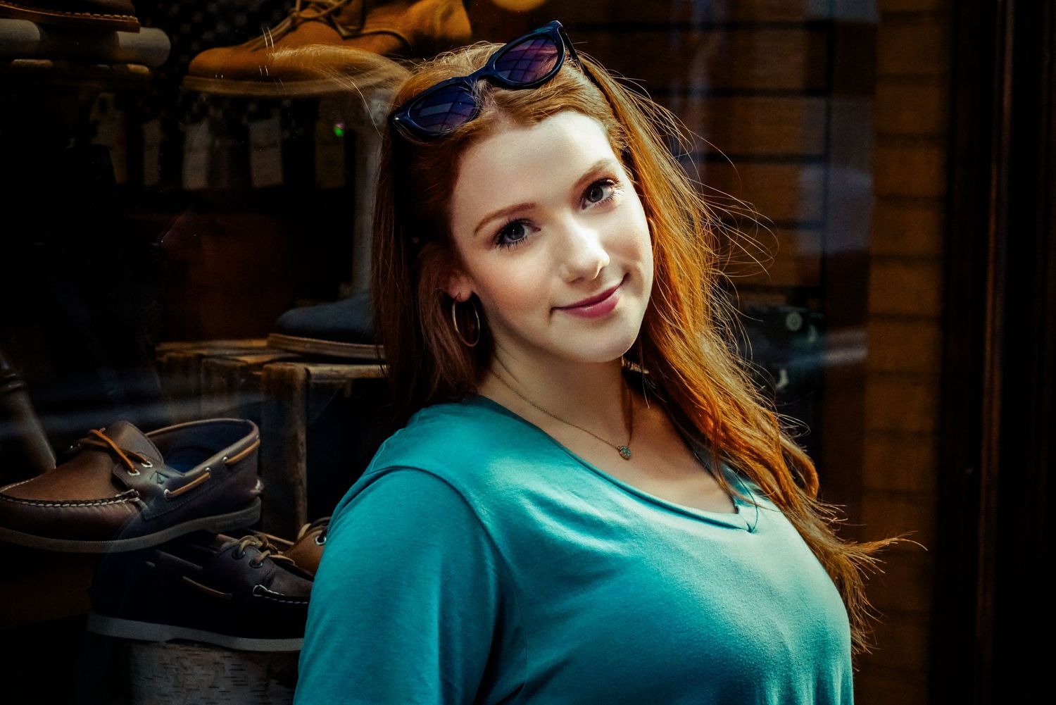 Red-haired woman in teal top smiling outdoors near a shoe store window display.