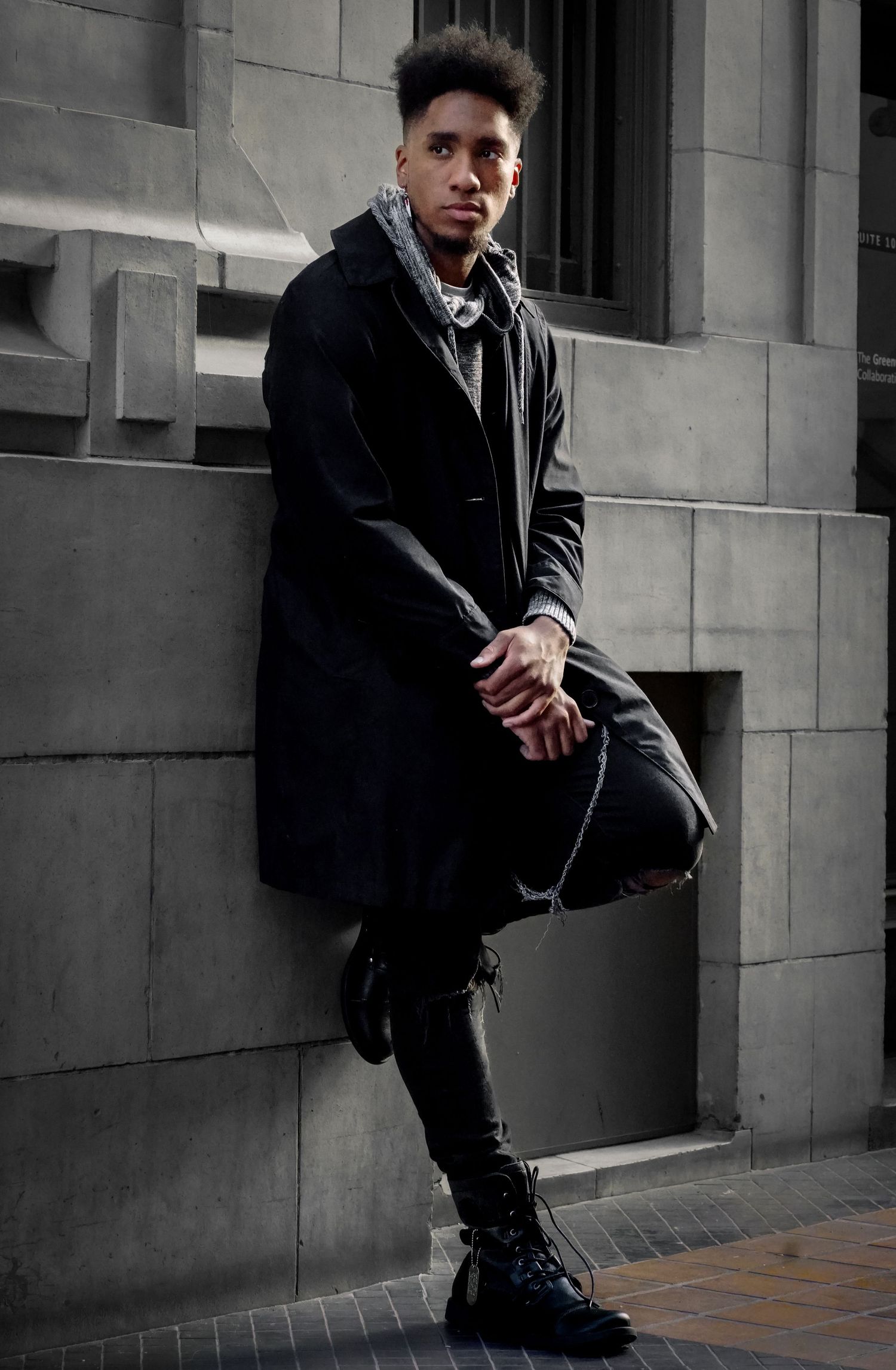 Young man in black coat and ripped jeans leaning against a stone wall outdoors in an urban setting.