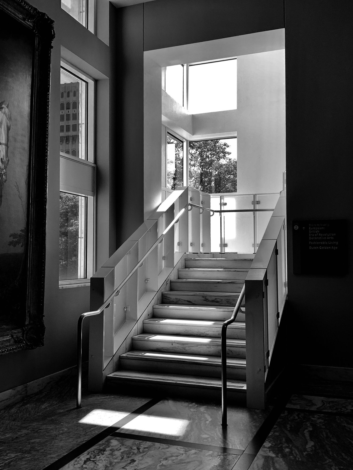 Black and white interior photo of elegant staircase with glass railings, marble steps, and natural light from large windows.