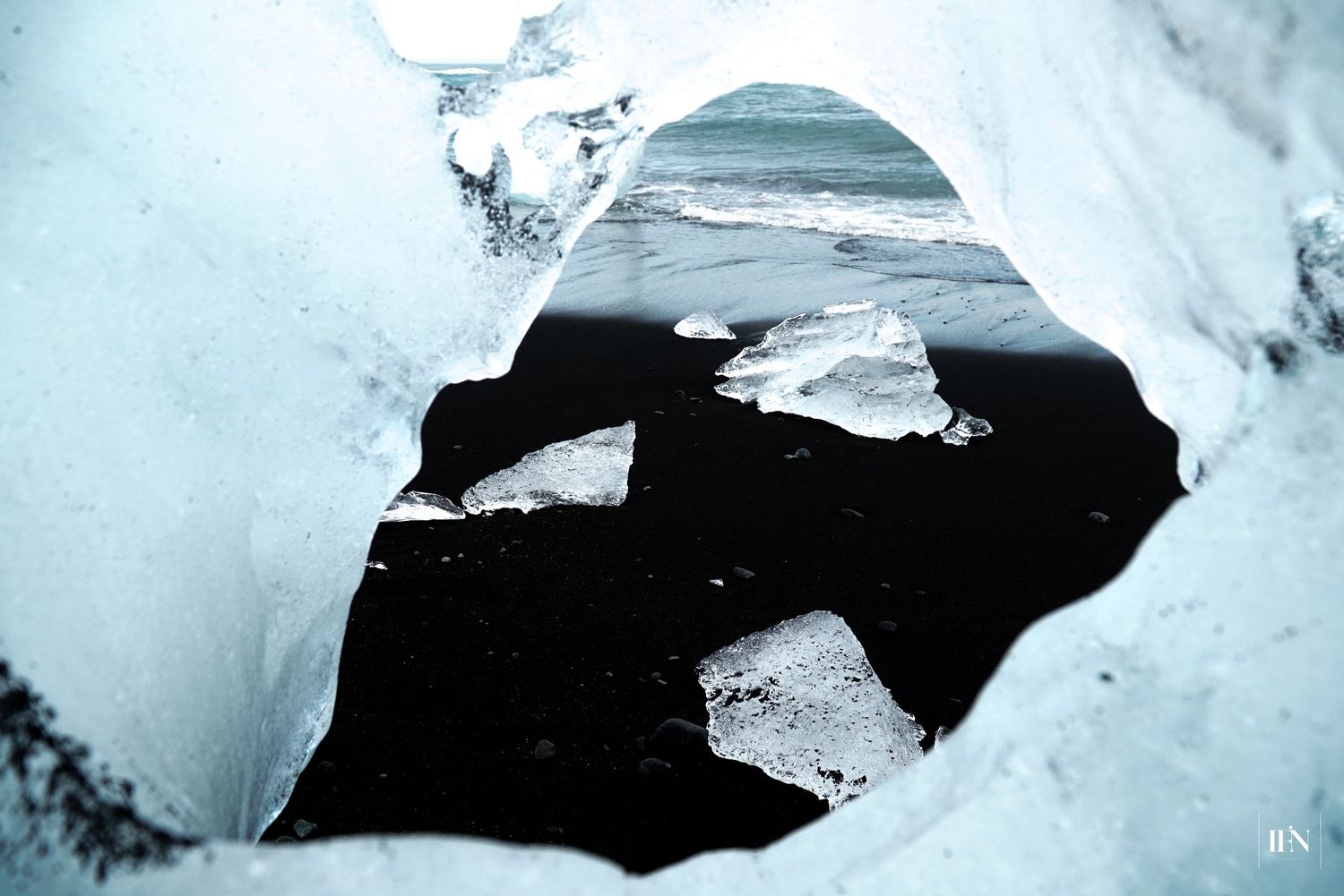 View through an ice cave opening revealing dark volcanic beach with scattered ice chunks in Iceland.