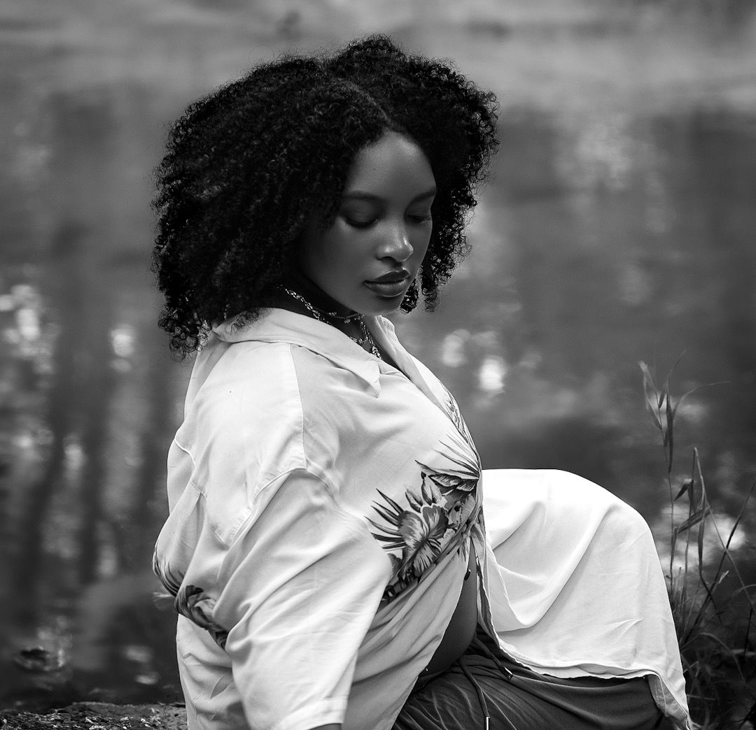 Black and white maternity portrait of a young woman with curly hair wearing a white shirt, sitting outdoors near water.
