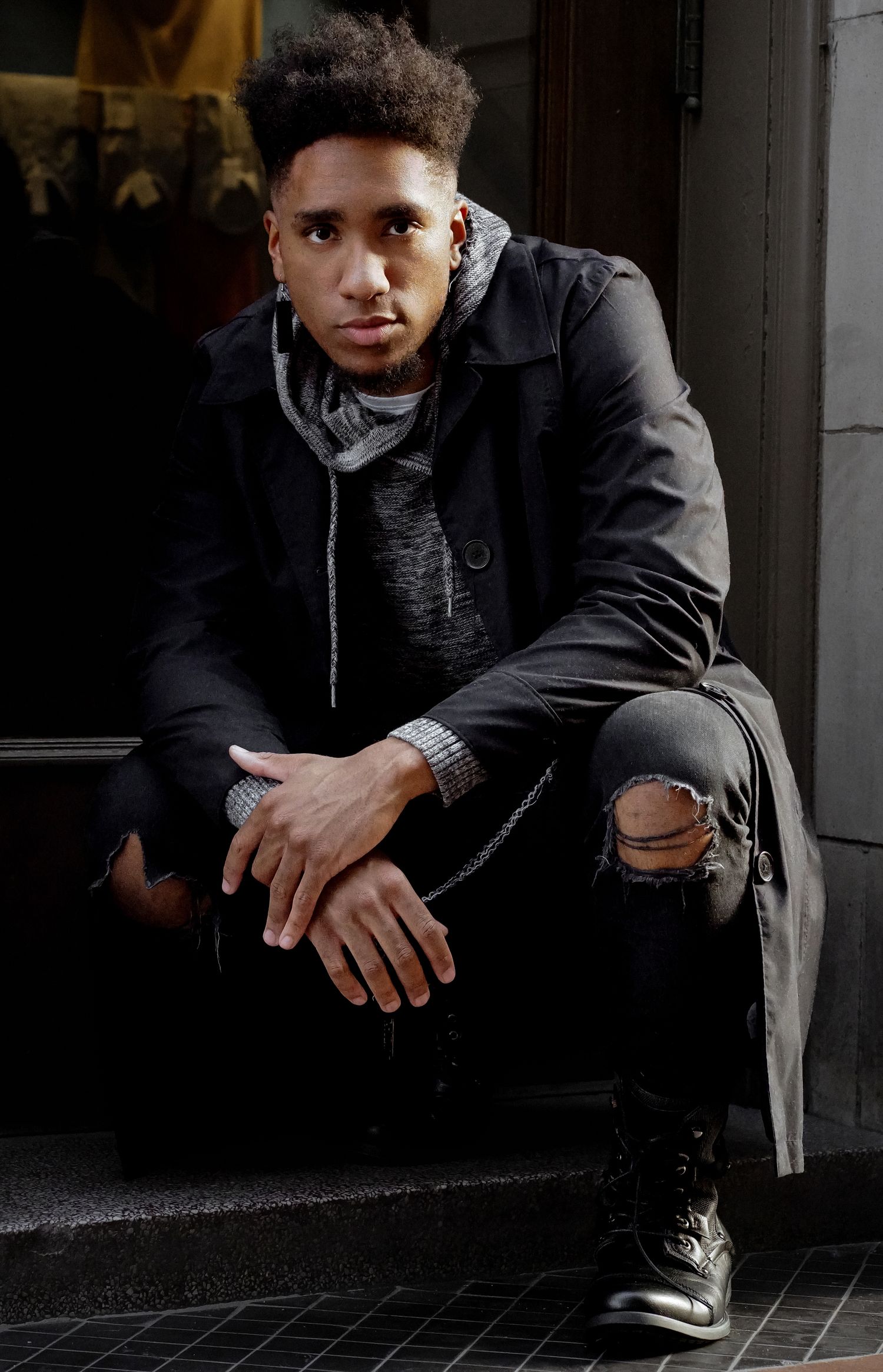 Young man in black ripped jeans and coat sits on steps, posing confidently with hands clasped.